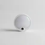 Veilleuse LED ronde et blanche avec petit capteur noir, vue de face sur fond gris clair