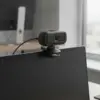 Webcam fixée par sa pince sur le haut de l'écran d'un ordinateur portable, dans un bureau lumineux